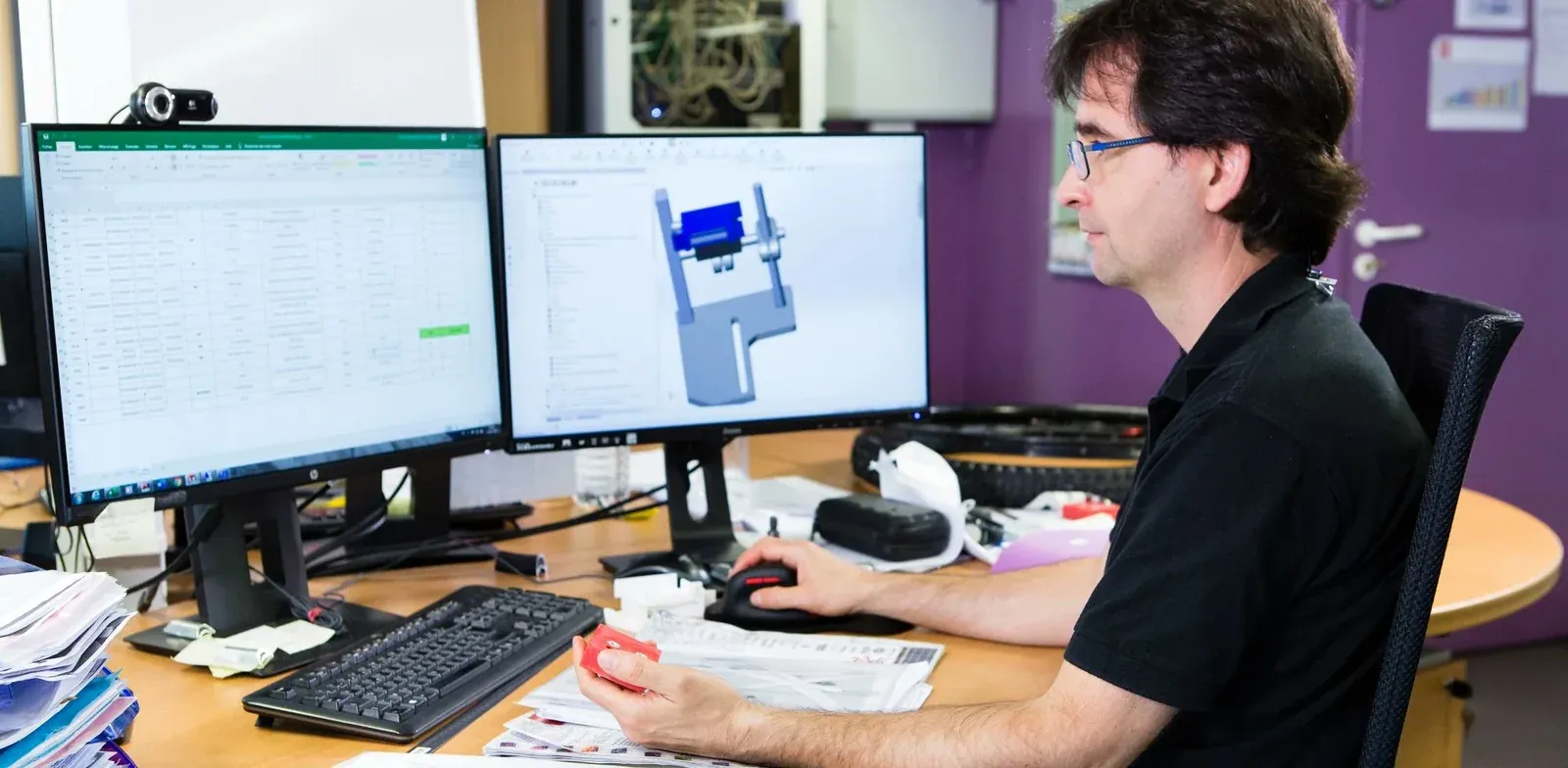 Un ingegnere dell'ufficio tecnico Mach1 davanti a due schermi, un foglio di calcolo e un disegno CAD, con in mano un pezzo stampato in 3D